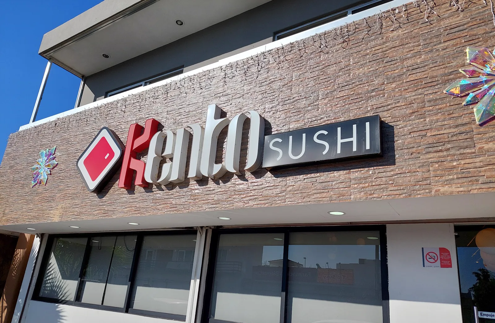 Fachada de Kento Sushi: su letrero con el rombo rojo sobre un muro de piedra.