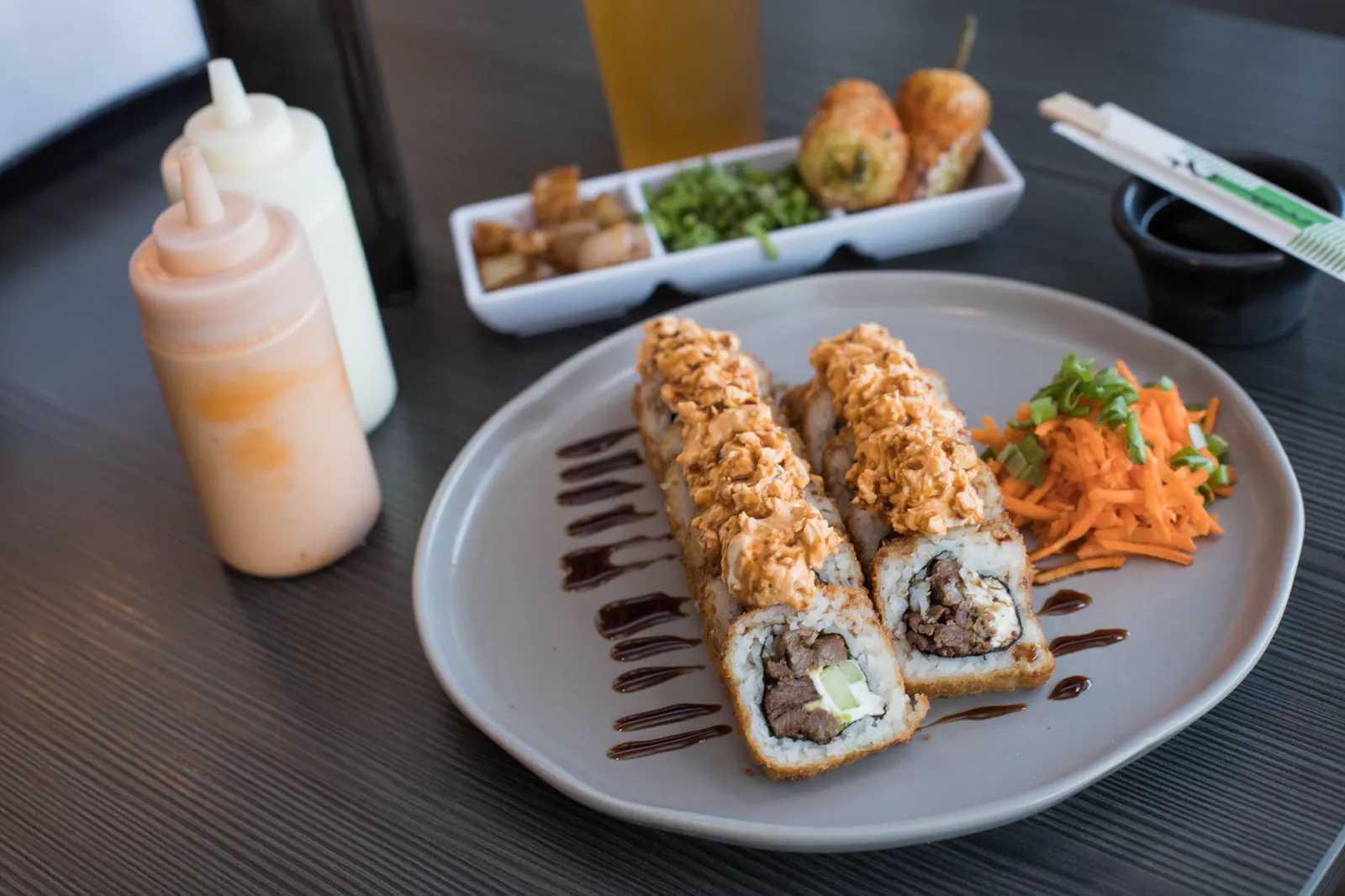 Rollos empanizados sobre plato gris con zanahoria rallada; al fondo, botellas de aderezo y otros platillos sobre la mesa oscura.